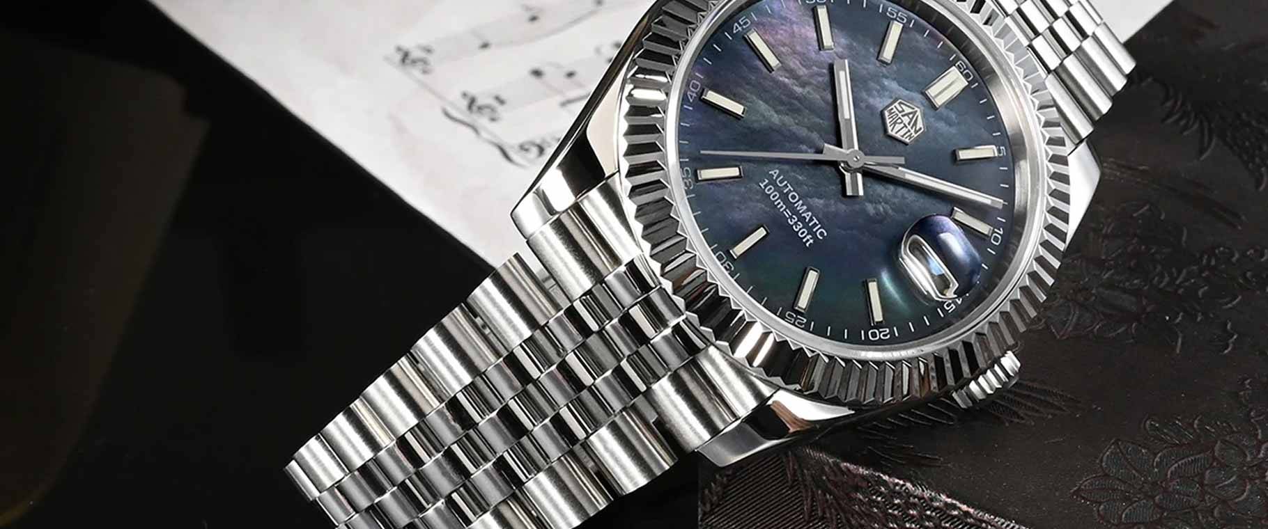 San Martin SN0058 Retro Homage Dress Watch featuring a stainless steel case and bracelet with a fluted bezel. The watch has a black mother-of-pearl dial with silver hour markers and hands, a date window with a magnifier, and is automatic with 100m water resistance. Displayed in a close-up, showcasing its luxurious and refined design.
