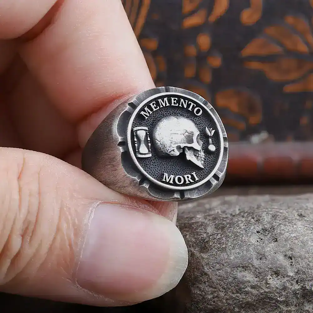 Close-up of a metallic Memento Mori ring with skull motif and engraved lettering.