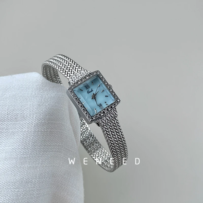 Luxury silver women's wristwatch with a rectangular face, gemstone accents, and woven mesh band.